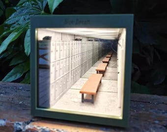 Może przedstawiać: Miniaturowa diorama korytarza z szafkami i ławkami. Scena zamknięta w zielonej ramie, z napisem "Blue Dream" u góry. Ławki są brązowe, a szafki w jasnoszarym kolorze.
