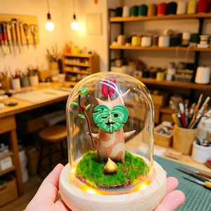 Puede incluir: Figurita artesanal de un espíritu del bosque, con una cara de hoja verde y un cuerpo de árbol, encerrada en una cúpula de cristal. La figurita se encuentra sobre musgo verde, sobre una base de madera con una luz cálida y brillante.