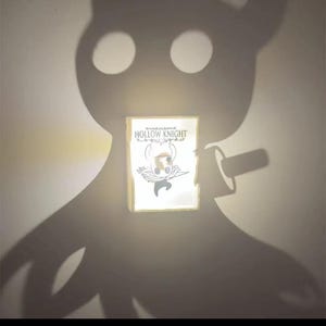 Könnte beinhalten: Ein leuchtender rechteckiger Leuchtkasten mit dem Logo und der Charakterkunst des Spiels "Hollow Knight". Der Leuchtkasten wird im Schatten einer Charakter-Silhouette gehalten, mit zwei großen augenartigen Kreisen darüber. Der Gesamteffekt erzeugt eine dramatische, beleuchtete Darstellung.