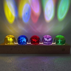 Puede incluir: Cinco pequeñas esferas de cristal translúcido iluminadas en amarillo, azul, rojo, morado y verde, sobre una base rectangular de madera. Cada esfera presenta un diseño grabado y texto único: Vah Naboris, Vah Ruta, Vah Rudania, Goddess Crest y Vah Medoh.