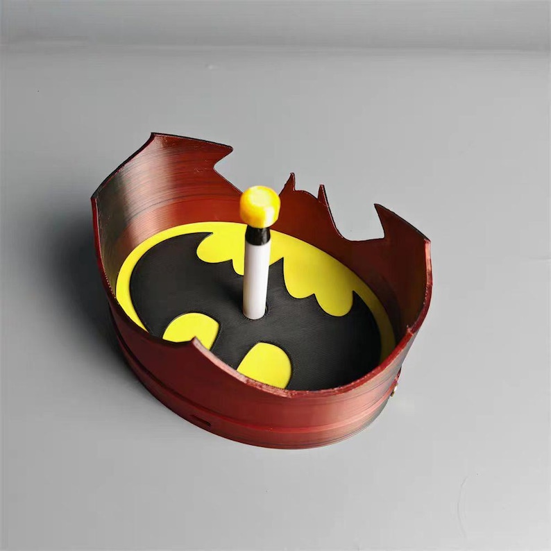 K&ouml;nnte beinhalten: Eine rot-schwarze Batman-Tablett mit einem gelb-schwarzen Batman-Logo in der Mitte. Ein wei&szlig;-schwarzes Objekt mit gelbem Oberteil befindet sich in der Mitte des Tabletts. Das Tablett hat einen fledermausf&ouml;rmigen Rand.