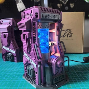May include: A purple, futuristic-looking device with a glowing blue interior. The device has a complex, mechanical design with visible text that reads "MVGS-055-YC" and "WAGS-022AC". The overall aesthetic suggests a science fiction or industrial theme.