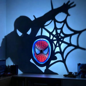 Può includere: Una lampada a forma di maschera di Spider-Man rossa, blu e bianca, con un bordo al neon blu. La luce proietta un'ombra di Spider-Man e una ragnatela sulla parete. La lampada è montata su una parete sopra una mensola di legno.