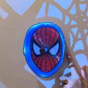 K&ouml;nnte beinhalten: Eine Spiderman-Maske mit rotem und schwarzem Spinnennetz-Design, umgeben von einem blau leuchtenden Rahmen. Die Maske hat wei&szlig;e Augenausschnitte und ein schwarzes kreisf&ouml;rmiges Nasenst&uuml;ck. Der Hintergrund hat ein Spinnennetz-Muster.