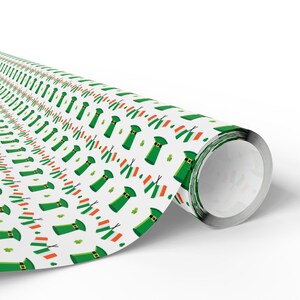 Könnte beinhalten: Eine Rolle weißes Geschenkpapier mit einem sich wiederholenden Muster aus grünen Koboldhüten, Kleeblättern und irischen Flaggen. Das Papier ist aufgerollt und zeigt das Muster auf der äußeren Schicht.