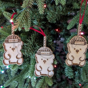 Puede incluir: Tres adornos navideños de madera con forma de oso colgados de un árbol de Navidad. Cada adorno tiene un nombre grabado: "Kenneth", "Jennifer Bear" y "Corina Bear". El año "2025" también está grabado. Los adornos están sujetos a cintas rojas.