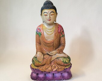 Op de afbeelding: Zittende Boeddha-beeld met een serene uitdrukking. Het beeld heeft een zwarte knot, gouden ketting en een oranje gewaad met bloemaccenten. Het staat op een paarse lotusbloem basis, met ingewikkelde details en een vredige uitstraling.