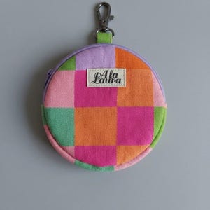 Puede incluir: Un monedero redondo de tela con un patrón de cuadros en tonos rosa, naranja y verde. El monedero tiene una cremallera color lila, un mosquetón de metal en la parte superior y una etiqueta rectangular blanca con el texto Ala Laura en el frente.
