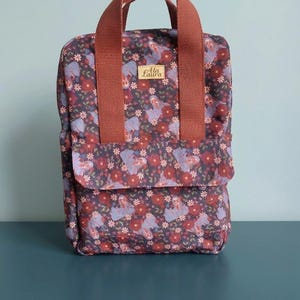 Può includere: Uno zaino rettangolare con un motivo floreale scuro di fiori rossi e viola su sfondo scuro. Presenta due spessi manici in tessuto color ruggine sulla parte superiore e una tasca frontale con patta. Un'etichetta riporta la scritta Ala Laura.