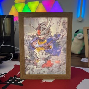 Puede incluir: Ilustraci&oacute;n enmarcada de un robot Gundam, con un dise&ntilde;o detallado en azul, rojo, amarillo y blanco. La obra de arte est&aacute; en un marco de madera marr&oacute;n claro, adecuado para su exhibici&oacute;n. La imagen est&aacute; iluminada desde atr&aacute;s.