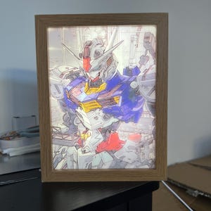 Puede incluir: Ilustraci&oacute;n enmarcada de un robot con una combinaci&oacute;n de colores azul, rojo, amarillo y blanco. El robot est&aacute; representado en una pose din&aacute;mica, con componentes mec&aacute;nicos detallados. El marco es de madera marr&oacute;n claro y la imagen est&aacute; iluminada desde atr&aacute;s.