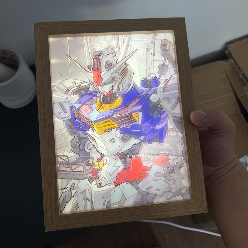 Puede incluir: Una obra de arte enmarcada e iluminada que presenta una ilustraci&oacute;n detallada de un robot. El robot es predominantemente blanco, azul, rojo y amarillo, sobre un fondo detallado. El marco es de madera marr&oacute;n claro.