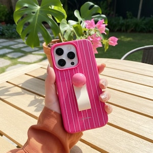 Pode incluir: Uma capa de telefone rosa brilhante com listras brancas verticais. A capa tem uma pega redonda rosa e uma alça branca. A capa do telefone é segurada em frente a uma mesa de madeira com uma planta ao fundo.
