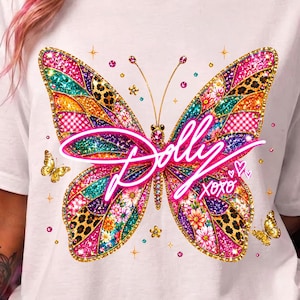 Op de afbeelding: Een wit t-shirt met een grote, kleurrijke vlindergrafiek met een mozaïekpatroon van luipaardprint, bloemen en ruitjes in roze, goud en groenblauw. De tekst "Dolly" en "xoxo" is gedrukt in een felroze cursief lettertype.