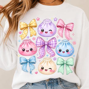 May include: A white long-sleeve sweatshirt featuring a colorful graphic print of four smiling, cartoon-style dumplings in purple, pink, blue, and cream, interspersed with five decorative bows in yellow, pink, purple plaid, blue, and green.