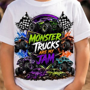 May include: A white short-sleeve t-shirt featuring a colorful graphic print of multiple monster trucks, checkered flags, and paint splatters. The text reads MONSTER TRUCKS ARE MY JAM in bold green, white, and purple lettering across the center.