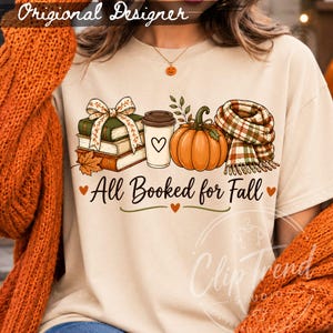 All Booked for Fall book lover design displayed on a cream T-shirt with books, pumpkin, coffee and plaid scarf in a Halloween fall setting.
