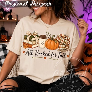 Cozy fall book lover shirt with All Booked for Fall quote, pumpkin, stacked books, coffee cup and plaid scarf styled with an orange sweater.