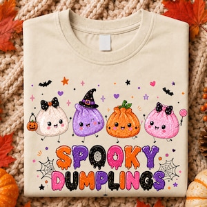 May include: A cream-colored t-shirt featuring a graphic of four cartoon dumplings with faces, including one wearing a witch hat, surrounded by bats, stars, and hearts. The text "SPOOKY DUMPLINGS" appears in a colorful, dripping font below.