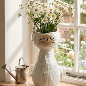 May include: A white ceramic cat-shaped vase filled with white daisies with yellow centers. The vase has a textured surface and black facial details. A small metal watering can and scissors sit nearby on a wooden windowsill.
