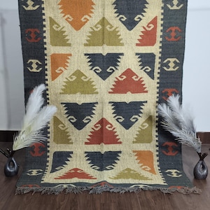Könnte beinhalten: Rechteckiger Teppich mit einem beigen Mittelfeld mit sich wiederholenden geometrischen Mustern in Orange, Grün, Marineblau und Rot. Der Teppich hat einen dunkelgrauen Rand mit dekorativen Elementen und einen Fransenrand. Der Teppich wird vor einer weißen Wand präsentiert.
