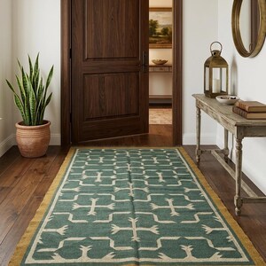 May include: A rectangular area rug featuring a deep teal green background with a repeating cream-colored geometric pattern. The rug has a solid mustard yellow border and dark brown fringe along the short ends, laid flat on a dark wood floor.