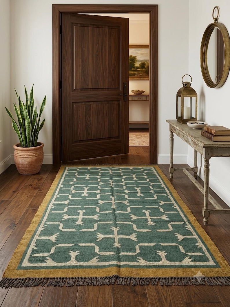 May include: A rectangular area rug featuring a deep teal green background with a repeating cream-colored geometric pattern. The rug has a solid mustard yellow border and dark brown fringe along the short ends, laid flat on a dark wood floor.
