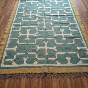 May include: A rectangular area rug featuring a teal green field with a repeating cream-colored geometric cross pattern. The rug is bordered by a solid mustard yellow band and finished with short, dark-toned fringe along the top and bottom edges.