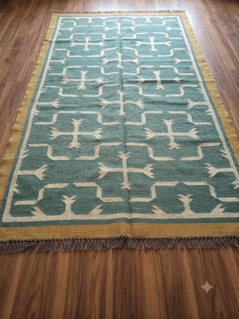 May include: A rectangular area rug featuring a teal green field with a repeating cream-colored geometric cross pattern. The rug is bordered by a solid mustard yellow band and finished with short, dark-toned fringe along the top and bottom edges.