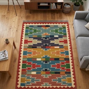 May include: A rectangular area rug featuring a repeating geometric pattern in shades of red, blue, green, yellow, and cream. The rug has a wide red border with a white sawtooth edge and short gray fringe along the top and bottom horizontal edges.
