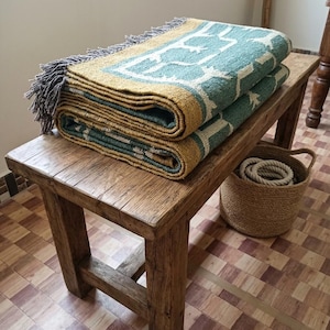 May include: A folded woven throw blanket featuring a teal and mustard yellow geometric pattern with grey fringe edges. The textile is layered on a wooden bench, showcasing a textured weave and a repeating symmetrical design across the fabric surface.