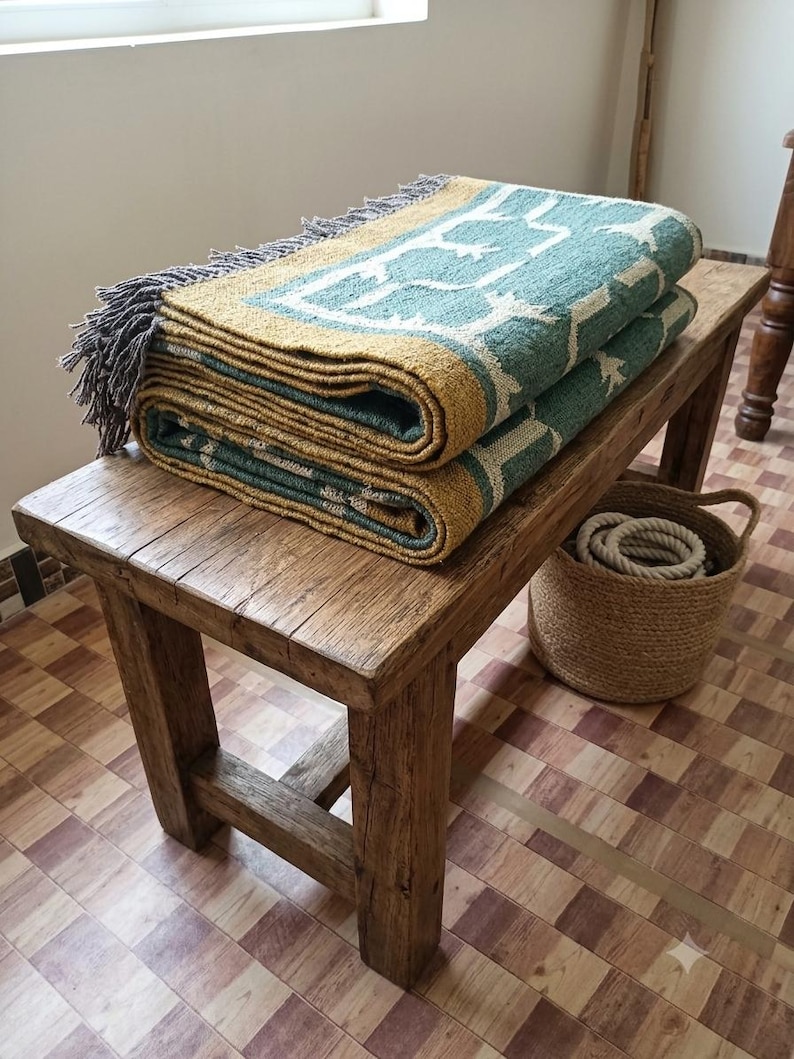 May include: A folded woven throw blanket featuring a teal and mustard yellow geometric pattern with grey fringe edges. The textile is layered on a wooden bench, showcasing a textured weave and a repeating symmetrical design across the fabric surface.