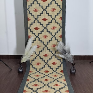 May include: A beige and grey geometric patterned runner rug with a dark grey border and fringe. The rug features a repeating pattern of dark grey diamonds with red accents. The rug is displayed vertically.