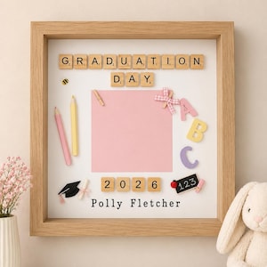 May include: A square wooden shadow box frame containing a white background with the text GRADUATION DAY and 2026 spelled out in wooden letter and number tiles. It features a pink square, pencils, ABC letters, a graduation cap, and the name Polly Fletcher.