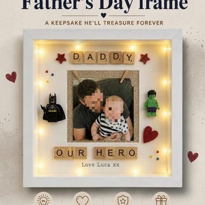 May include: A white framed Father's Day keepsake. The frame contains a photo of a father and child, the text "My Hero Father's Day frame", and the word "Daddy" in Scrabble tiles. It is decorated with a Batman figurine and fairy lights.