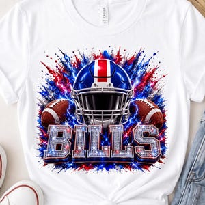 Puede incluir: Una camiseta blanca de manga corta con un estampado gráfico de un casco de fútbol americano azul, rojo y blanco flanqueado por dos balones. El diseño incluye salpicaduras azules y rojas y la palabra BILLS en una fuente plateada brillante.