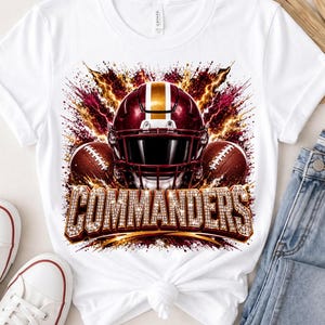 Może przedstawiać: Biały t-shirt z krótkim rękawem z nadrukiem graficznym przedstawiającym bordowo-złoty kask do futbolu amerykańskiego w otoczeniu dwóch piłek. Wzór zawiera efekty błyskawic oraz napis COMMANDERS wykonany teksturowaną czcionką przypominającą diamenty.
