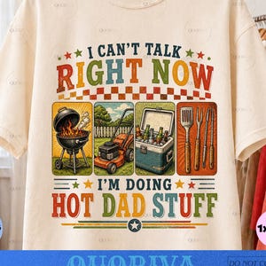 May include: A cream-colored short-sleeve t-shirt featuring the text I CAN'T TALK RIGHT NOW I'M DOING HOT DAD STUFF. The graphic includes illustrations of a charcoal grill, a lawnmower, a cooler filled with bottles, and various grilling utensils.
