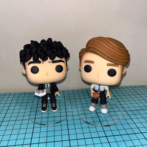 Puede incluir: Dos figuras de vinilo de personajes con grandes ojos negros. Una figura tiene cabello negro rizado, un traje negro y zapatos blancos y negros. La otra figura tiene cabello castaño, una camisa blanca y zapatos grises y blancos. Ambas figuras están sobre bases circulares transparentes.