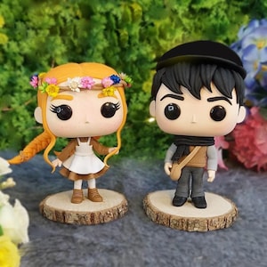 May include: Two Funko Pop-style figurines, one with red hair and a flower crown, wearing a brown coat and white dress. The other has black hair, a black cap, and a scarf. Both stand on wooden slices.