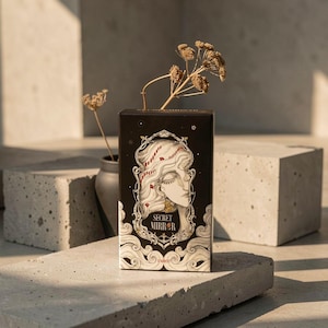 May include: A rectangular tarot deck box with a black background featuring an ornate gold-toned frame surrounding a stylised illustration of a person's face. The text "SECRET MIRROR" is printed in gold lettering, with "TAROT" below the frame.