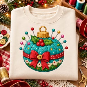 May include: A cream-colored crewneck sweatshirt featuring an embroidered design of a teal, round pincushion shaped like a Christmas ornament. The design includes a gold cap, a red bow with a button center, holly leaves, red berries, and colorful sewing pins.