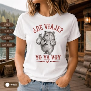 May include: White t-shirt featuring a detailed illustration of a hamster carrying a bundle and a cookie. The text "¿DE VIAJE? YO YA VOY" is printed in red above and below the graphic. The shirt is a casual style.