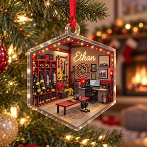 May include: A hexagonal acrylic ornament featuring a detailed fire station interior scene. The room includes red lockers with gear, a desk with a computer, a fire rescue emblem, and the name Ethan in white script. Red string lights line the top.