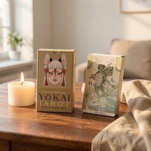 May include: Two tarot card decks on a wooden table. One deck features a fox-like face with red accents, and the text "YOKAI TAROT". The other card shows a turtle-like figure. A lit candle is to the left.