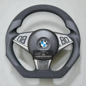 Pode incluir: Um volante BMW preto e cinza com design em fibra de carbono. O volante tem uma pega em couro perfurado, detalhes prateados e o logótipo BMW no centro. O texto "SA Carbographics Carbon Fiber Auto Parts" é visível.