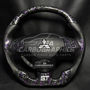 Könnte beinhalten: Ein schwarzes und lila Carbonfaser-Lenkrad mit perforiertem Ledergriff. Das Lenkrad hat ein Mitsubishi-Logo und den Text "SA Carbographics Carbon Fiber Auto Parts". Das Lenkrad hat ein GT-Emblem.
