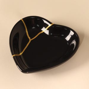 May include: A heart-shaped dish with a glossy black finish, featuring a prominent gold-colored line that runs across the surface, mimicking the appearance of a repaired break. The dish has smooth, curved edges and a reflective, polished surface.