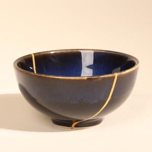 May include: A ceramic bowl featuring a deep blue and black gradient glaze with a prominent gold-colored repair line running from the rim down to the base, showcasing a kintsugi-style aesthetic with a smooth, glossy finish and a rounded shape.