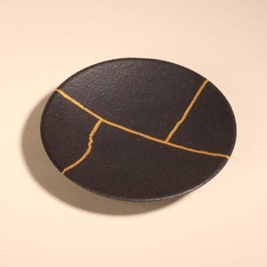 May include: A circular, black ceramic plate featuring gold-coloured kintsugi-style repair lines. The plate has a textured surface and a simple design. The gold lines highlight the cracks, creating a unique decorative piece.